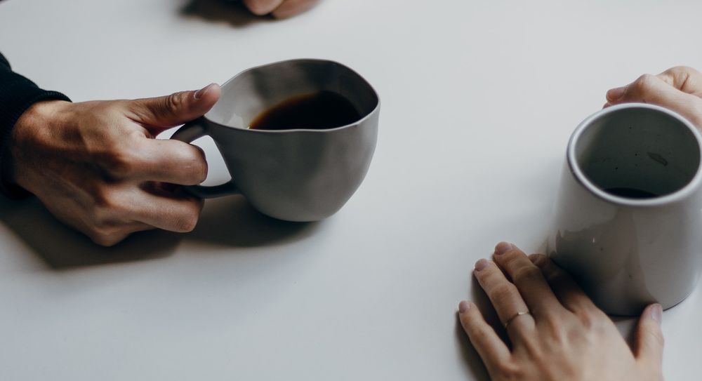 Two pairs of hands holding a coffee cup each, across from each other on a table. 