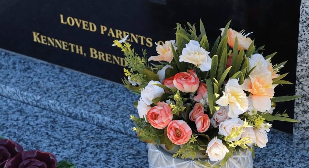 A vase of white, yellow and pink funeral flowers next to a black marble headstone