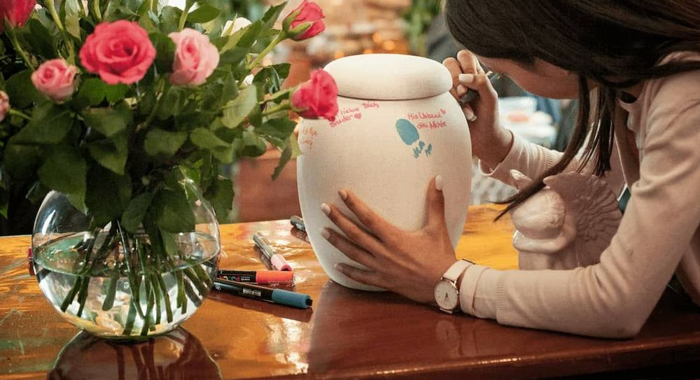 A woman writing on a plain white cremation urn with a paint marker