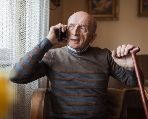 Senior man with walking stick having mobile phone conversation at home