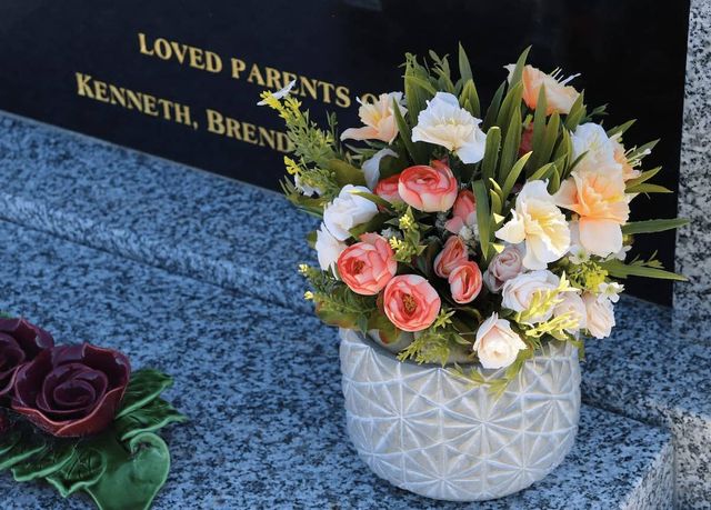 A vase of white, yellow and pink funeral flowers next to a black marble headstone