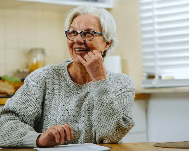 A senior woman sits at her kitchen table smiling, with paperwork in front of her