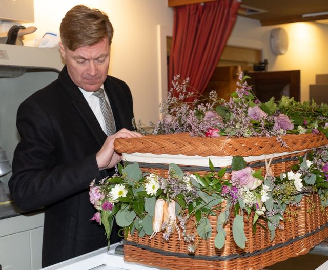 Man in black suit standing next to a wicker coffin decorated with flowers